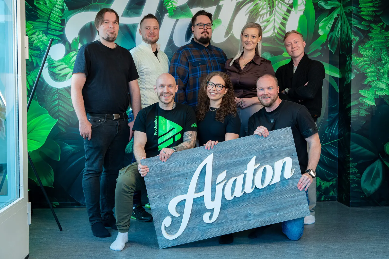 Ajaton Idean kahdeksan työntekijää ryhmäkuvassa viidakkotapetin edessä, edessä puinen Ajaton-kyltti.